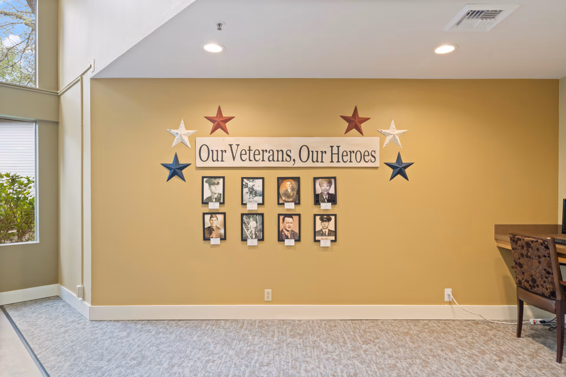 A beige interior wall display reading 'Our Veterans, Our Heroes' with framed photos and decorative stars in a senior living common area.