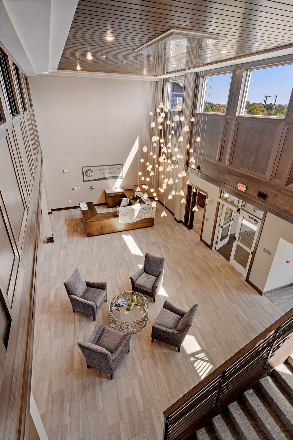 View from above of a spacious lobby area with a modern chandelier hanging from the ceiling, four gray armchairs arranged around a round glass table, a wooden reception desk with a sign that reads 'Meadowview of Clive,' large windows letting in natural light, and a staircase with a wooden handrail.