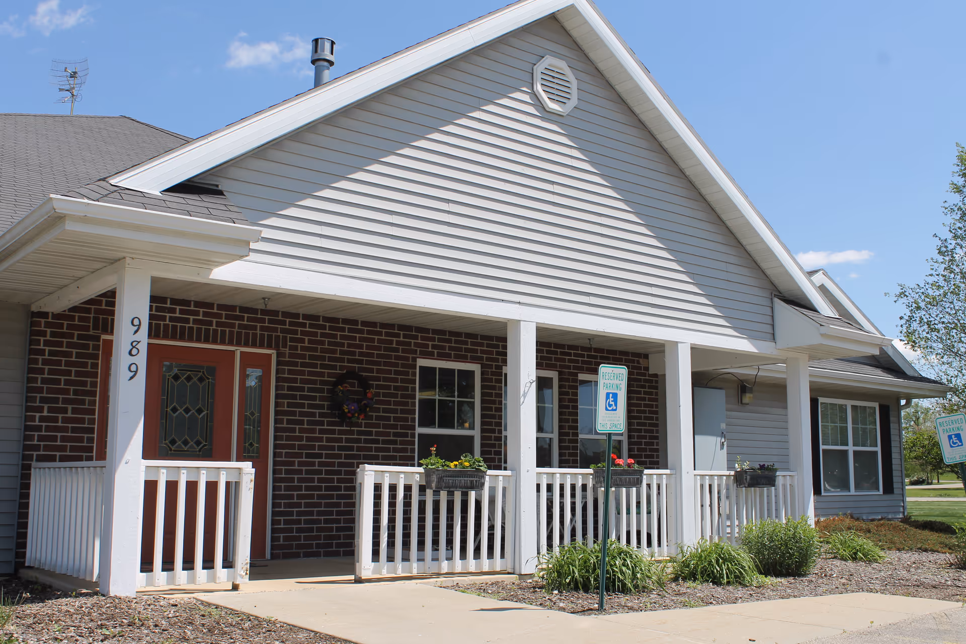 Front entrance of a single-story memory care building with a covered porch, white railing, and reserved handicap parking signs.
