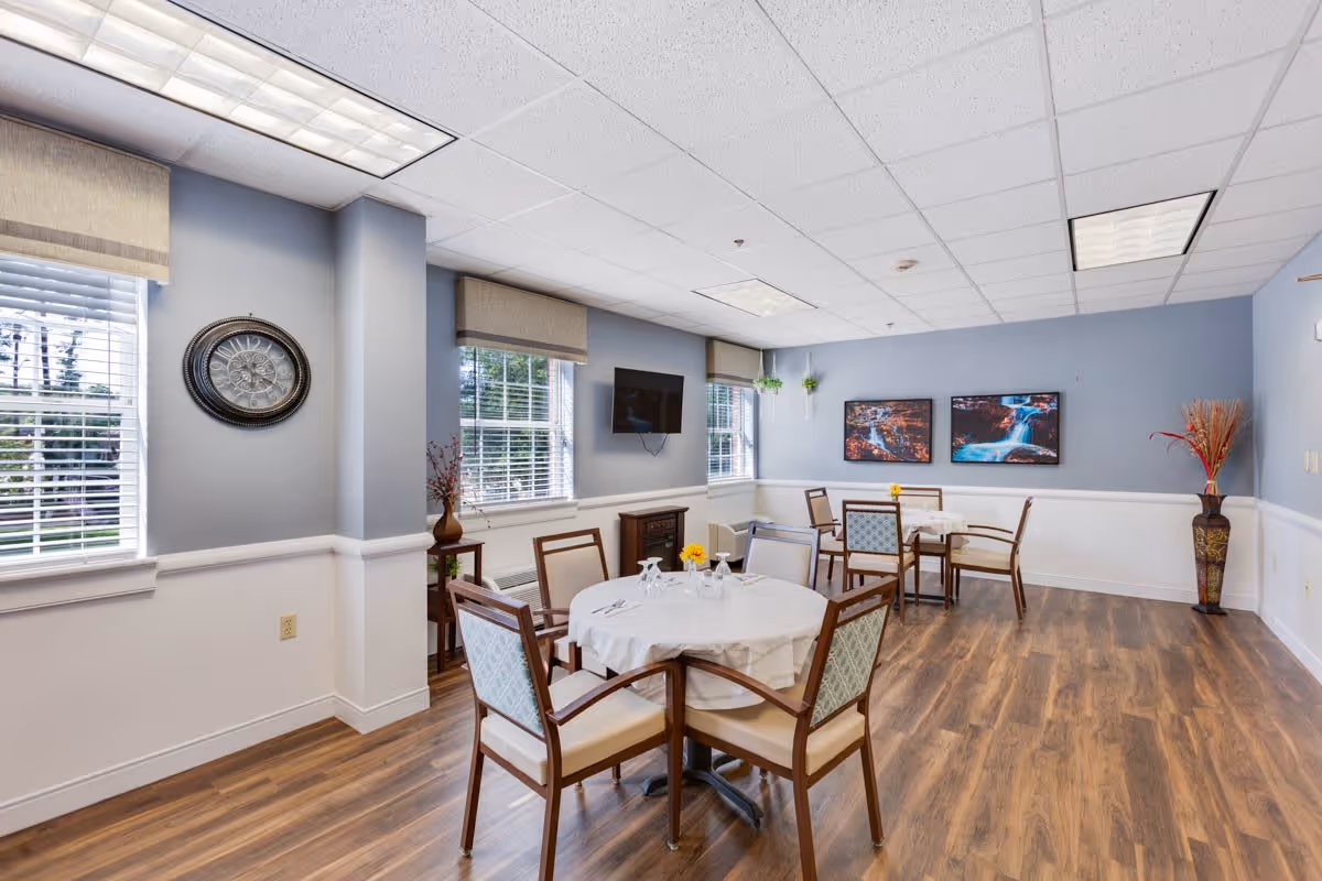 A bright dining room with light blue walls and wood flooring. The room has three round tables covered with white tablecloths, each surrounded by four wooden chairs with cushioned seats and backs. There are two large windows with blinds and beige valances, a wall-mounted TV, a large decorative clock, two framed pictures of waterfalls on the far wall, and a tall vase with dried plants in the corner.