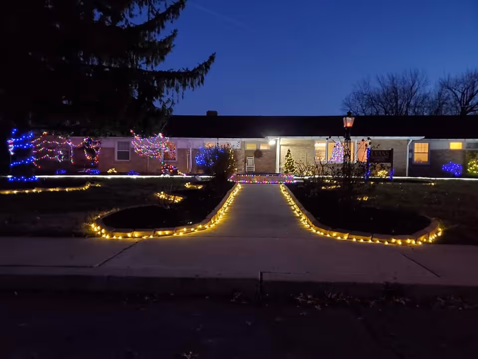 Nighttime view of the Evans Retirement Center building decorated with colorful holiday lights along the bushes, trees, and walkway edges. The building is single-story with lit windows and a sign near the entrance. The sky is dark blue, indicating early evening or night.