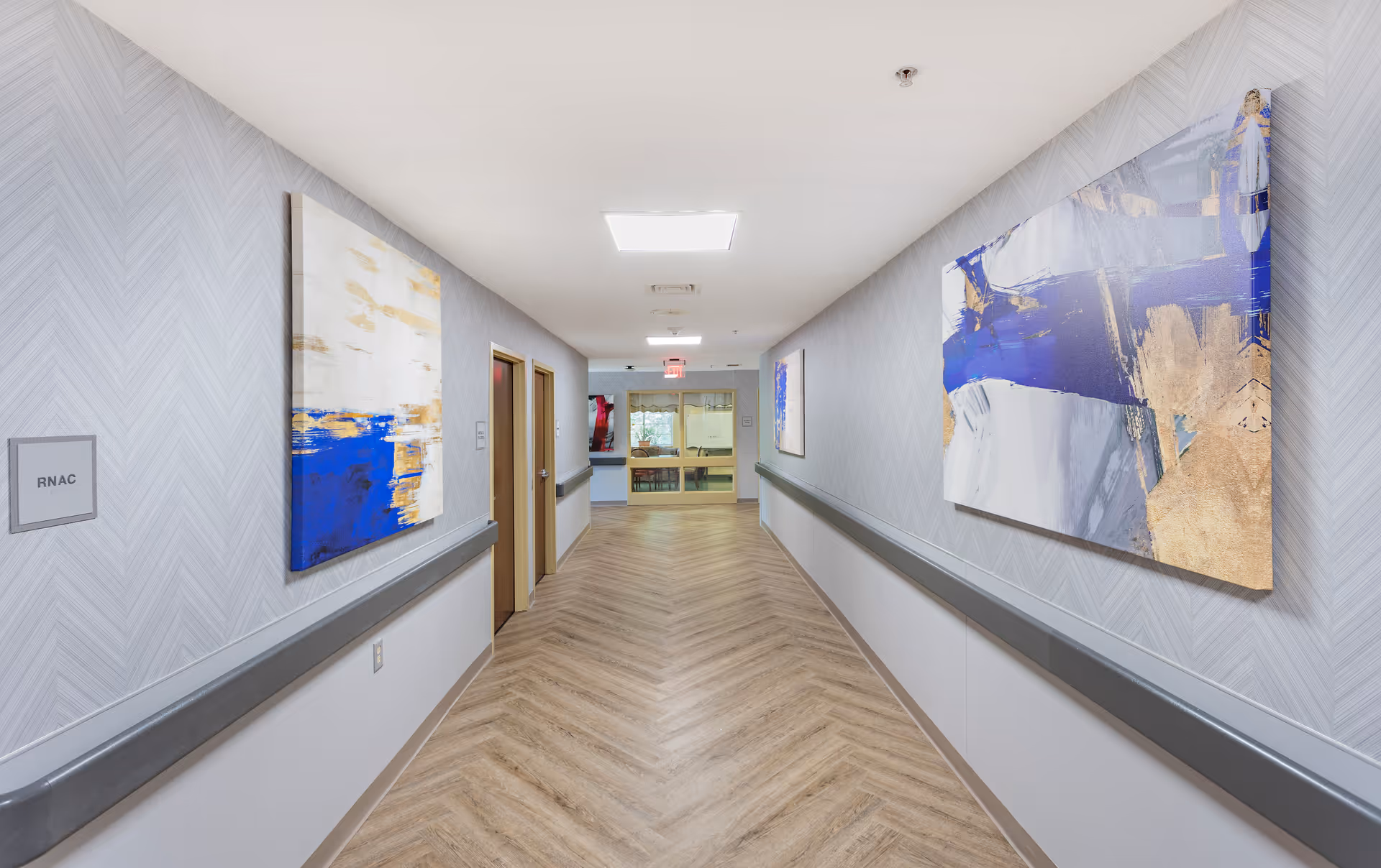 Bright, modern interior hallway with handrails, abstract paintings on the walls, wood-patterned flooring, and doors leading to rooms.