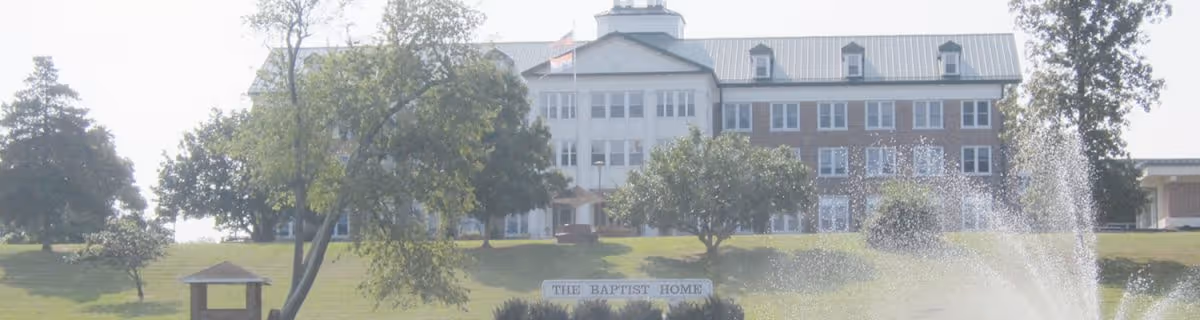 A large multi-story brick building with white trim and a metal roof, identified as The Baptist Home, situated on a grassy hill with several trees and a water fountain spraying in the foreground.