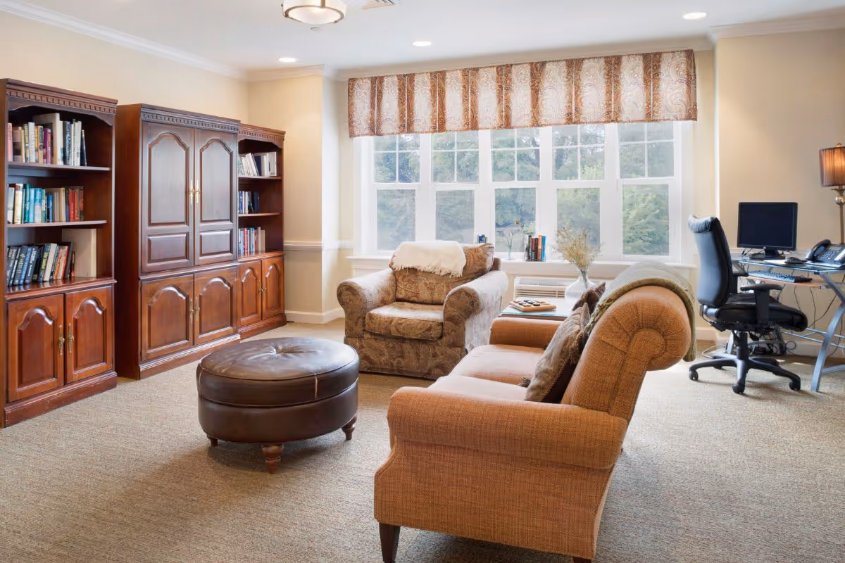 A cozy living room with a large window covered by a patterned valance, letting in natural light. The room features a brown upholstered armchair with a white throw draped over the back, a matching sofa with cushions, and a round leather ottoman in the center. Against one wall are wooden bookshelves filled with books. A desk with a computer and office chair is positioned near the window. The room has beige carpeting and warm-toned walls.