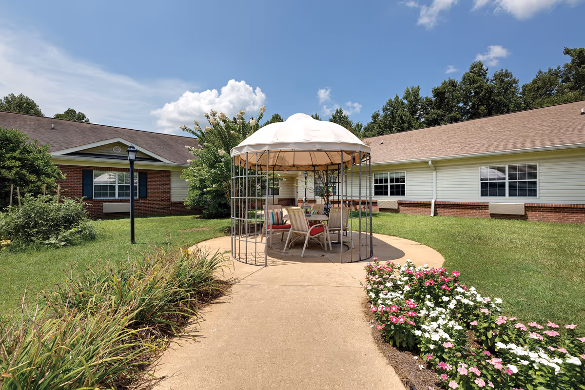 Outdoor courtyard area at The Pinnacle of Oxford featuring a circular paved seating area with a metal gazebo structure covered by a white canopy. There are several chairs with cushions around a table under the gazebo. The courtyard is surrounded by green grass, flowering plants, and a building with brick and siding exterior walls under a partly cloudy blue sky.