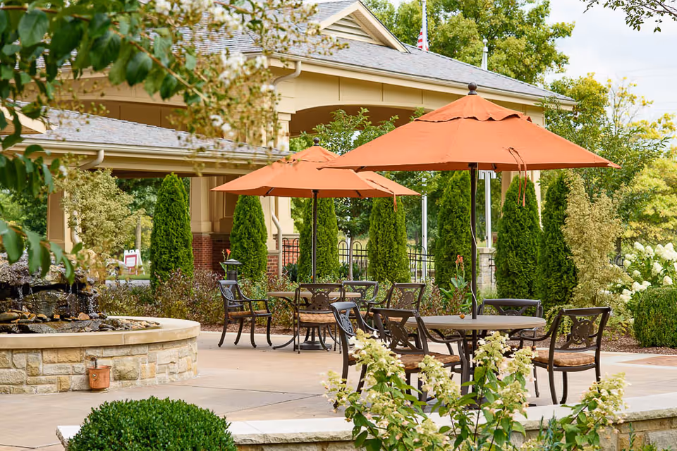 Outdoor patio area with round tables and metal chairs under orange umbrellas, surrounded by green shrubs and trees, with a stone water fountain on the left and a building with a covered porch in the background.
