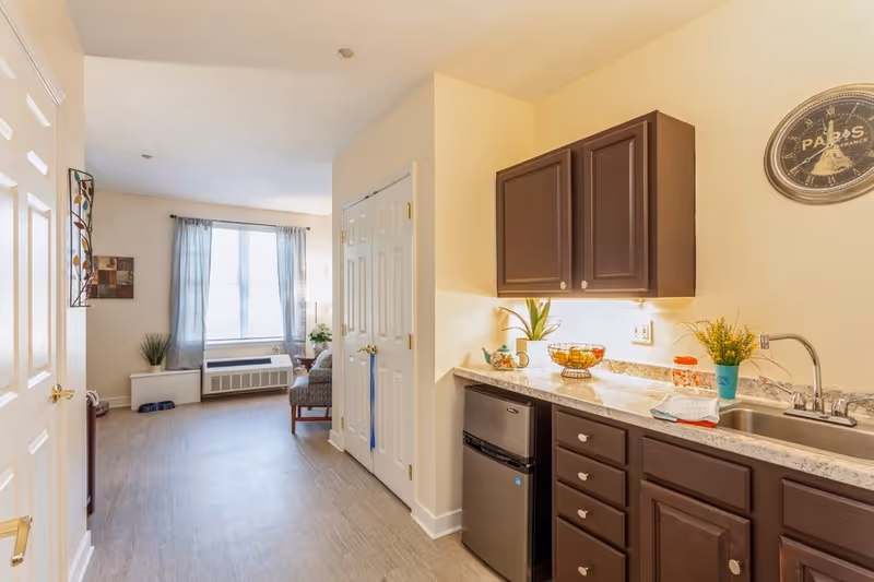 Interior view of a senior living facility showing a small kitchenette area with dark brown cabinets, a countertop with a sink, and a mini refrigerator. The kitchenette opens into a living area with a window covered by sheer curtains, a chair, and some decorative plants. The walls are painted light yellow, and there is a round wall clock above the sink.