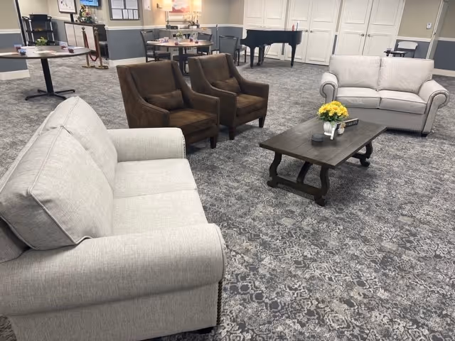 Communal lounge area with sofas, armchairs, a coffee table, dining tables and a piano in a senior living facility.