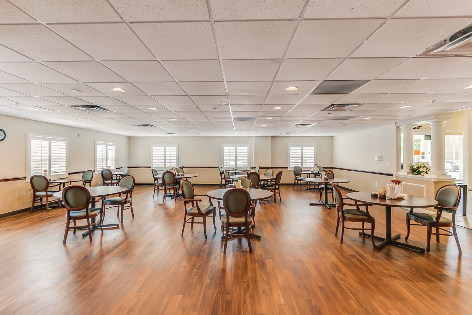 A spacious dining room with multiple round tables and green cushioned chairs arranged neatly on a wooden floor. The room has white walls, several windows with white shutters, and a ceiling with recessed lighting. There are small flower arrangements on some tables and a clock on the wall to the left.