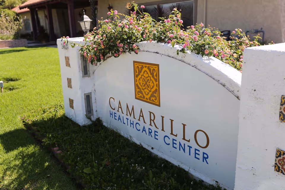 Outdoor white sign reading "Camarillo Healthcare Center" surrounded by flowering plants and a green lawn in front of the building.