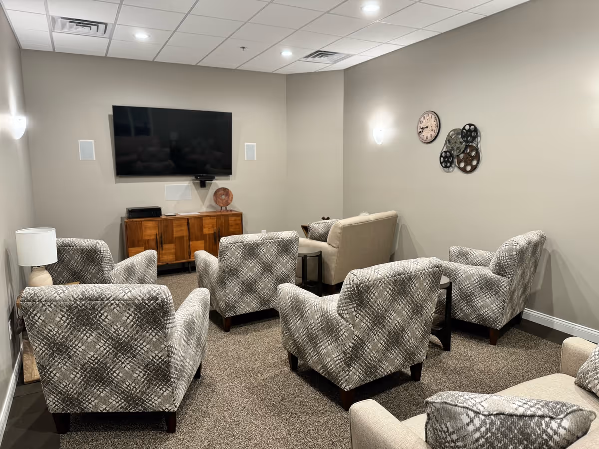 A cozy living room with six patterned armchairs and one beige loveseat arranged facing a wall-mounted flat-screen TV. The room has beige walls, carpeted floor, a wooden cabinet under the TV, a table lamp on a side table, a wall clock, and decorative film reels on the wall.