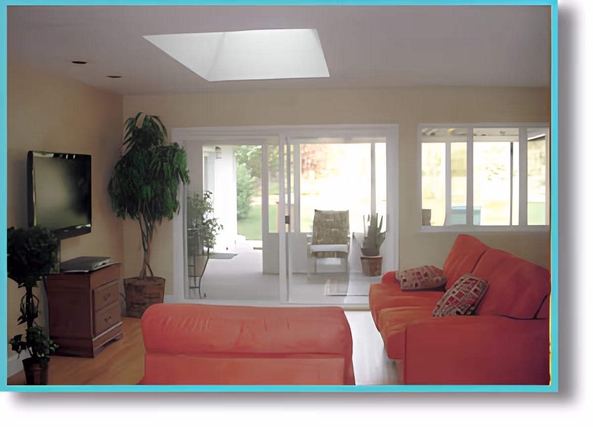 A living room with a red sofa and matching ottoman, two patterned cushions on the sofa, a flat-screen TV mounted on the wall, a wooden cabinet below the TV, several potted plants, and large sliding glass doors leading to a bright outdoor patio area.