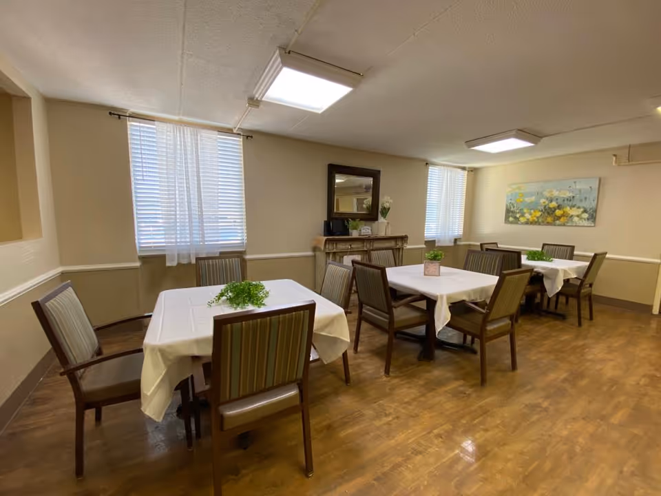 Small communal dining room with several tables covered in white tablecloths, chairs, potted plants, and wall art.