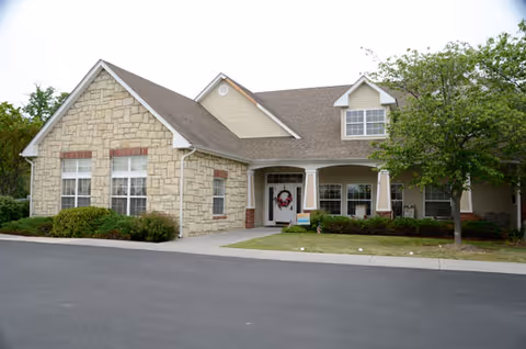 Exterior view of a single-story building with stone and siding facade, a covered entrance with columns, a wreath on the door, bushes and a tree in front, and a paved driveway.