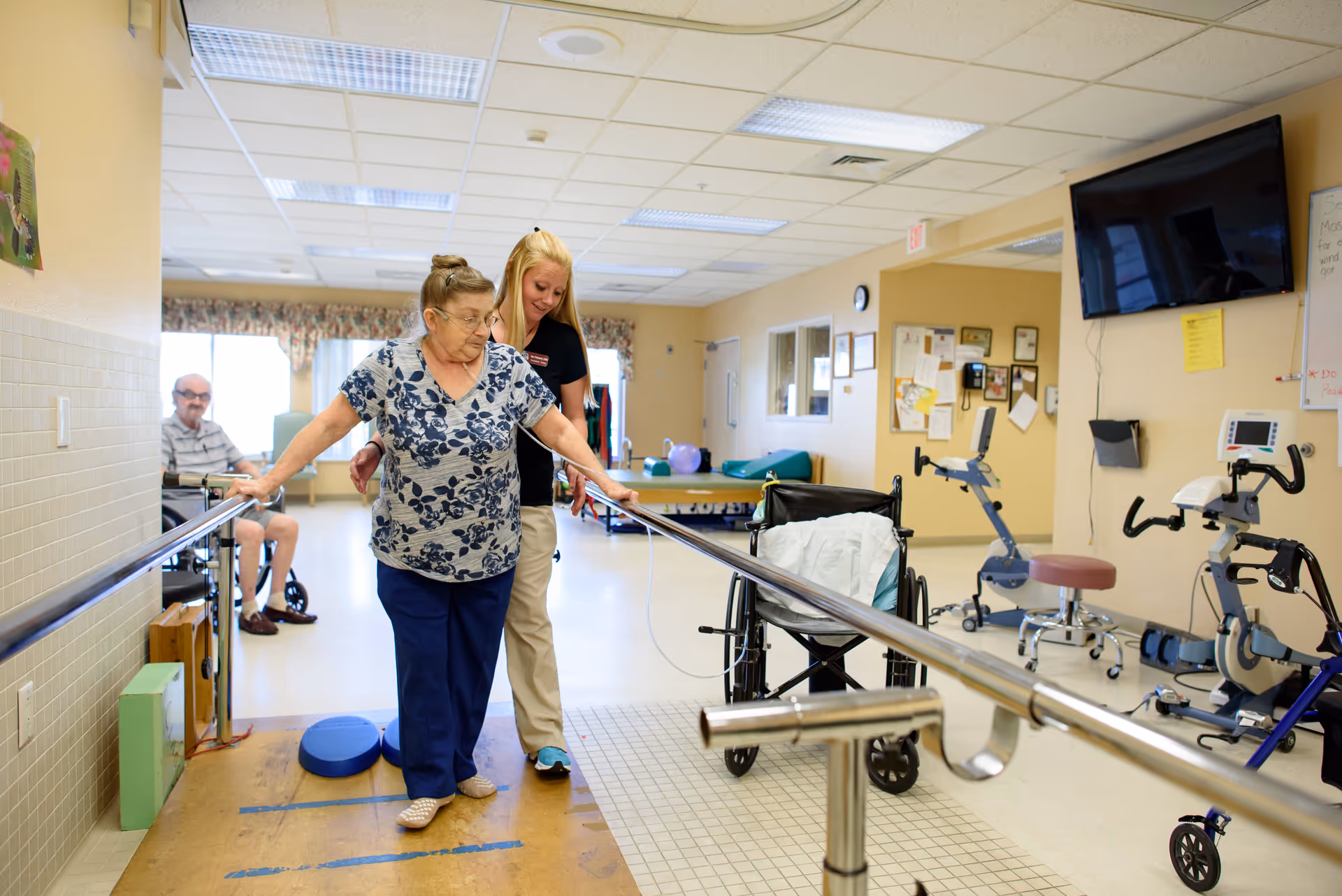 An elderly woman is walking with the assistance of parallel bars in a rehabilitation or physical therapy room, supported by a caregiver standing behind her. In the background, a man is sitting in a wheelchair. The room contains exercise equipment, a wheelchair, and a wall-mounted TV.