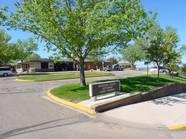 Driveway and landscaped front of Missouri Slope Lutheran Care Center with a low stone sign, trees, and a single-story building.