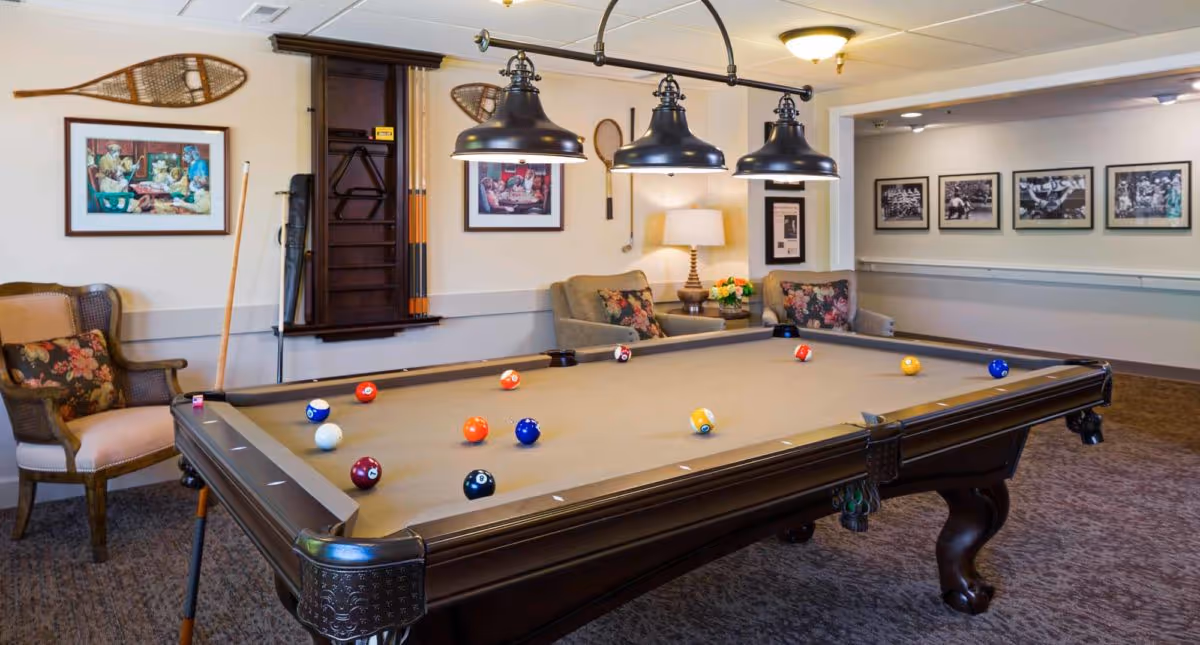 A cozy game room with a billiards table in the center, surrounded by comfortable armchairs with floral cushions. The walls are decorated with framed pictures and vintage tennis rackets. Overhead, three black pendant lights illuminate the pool table. The room has a warm and inviting atmosphere.