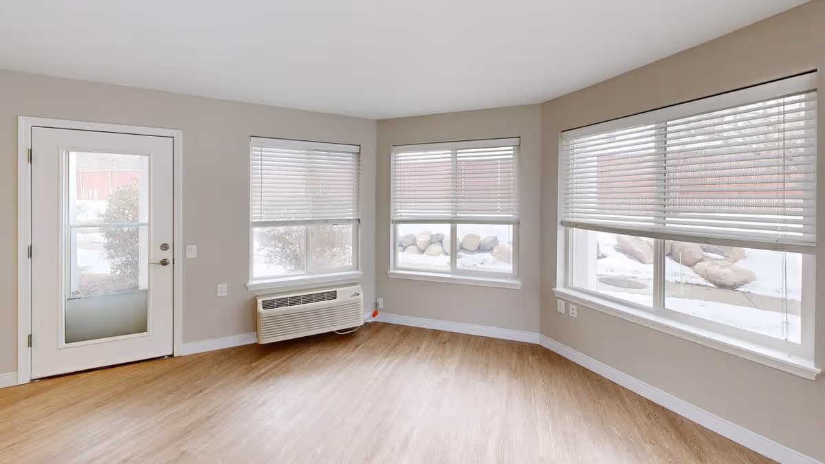 Empty room with light wood flooring, beige walls, three large windows with white blinds partially open, and a white door with a glass panel. Outside the windows, snow and rocks are visible.