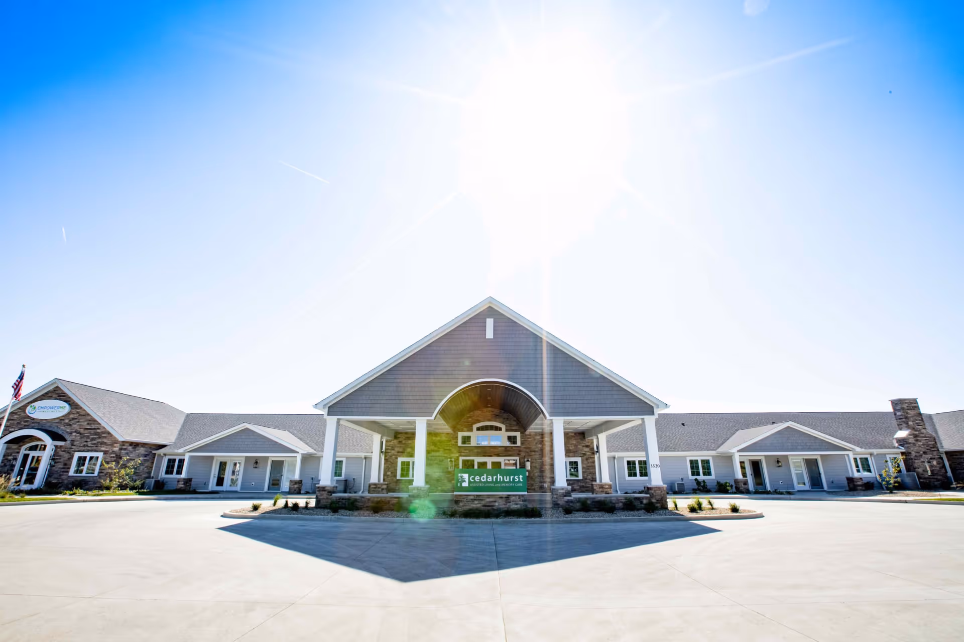 Front exterior view of Cedarhurst of Lebanon, a single-story assisted living and memory care facility with a large covered entrance, stone and siding facade, and clear blue sky with bright sunlight.