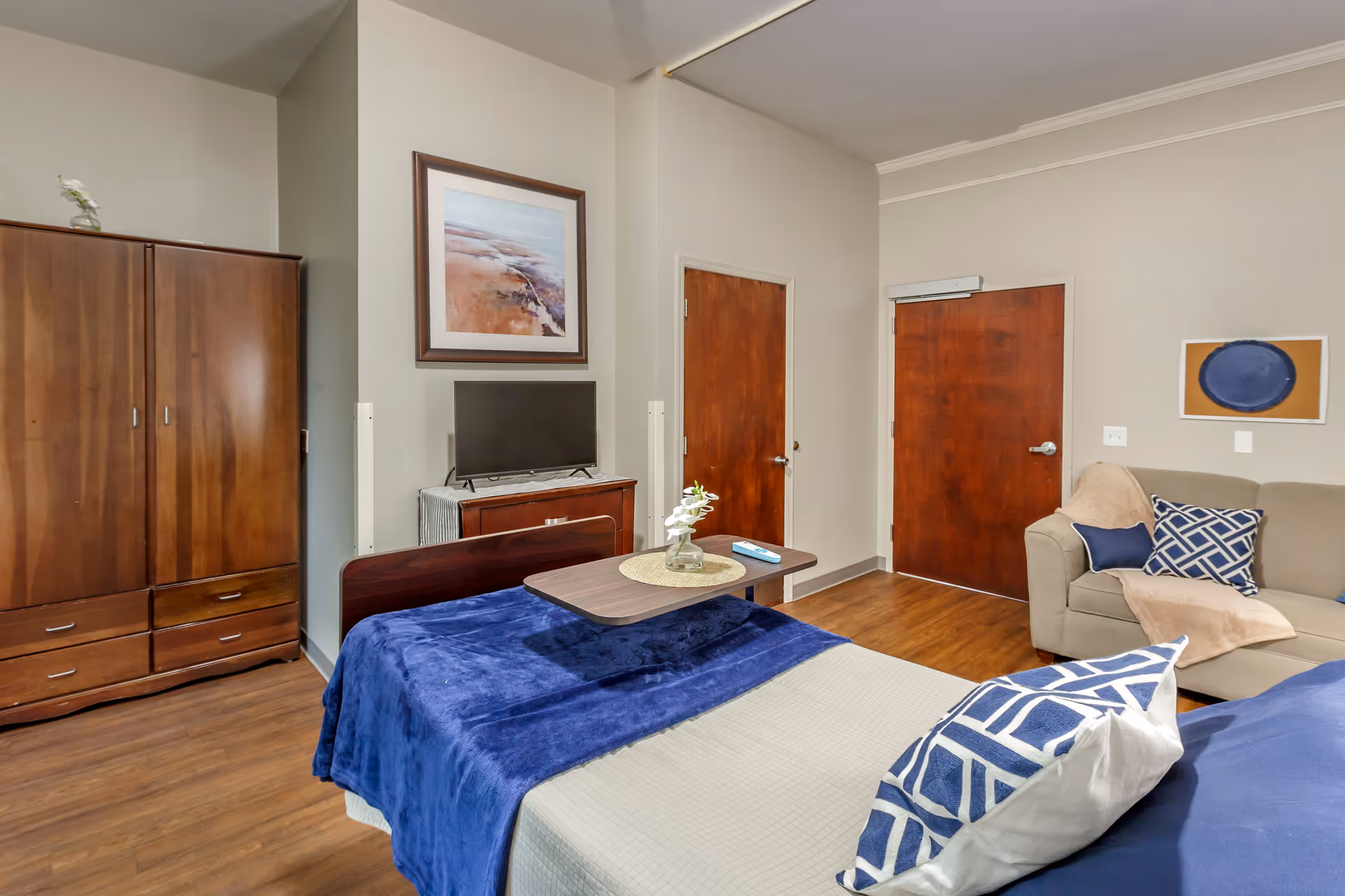 A senior living facility bedroom with a bed covered in blue and beige bedding, a wooden wardrobe, a small TV on a wooden dresser, a beige couch with blue and beige pillows, and two wooden doors. The room has wooden flooring and neutral-colored walls with framed artwork.
