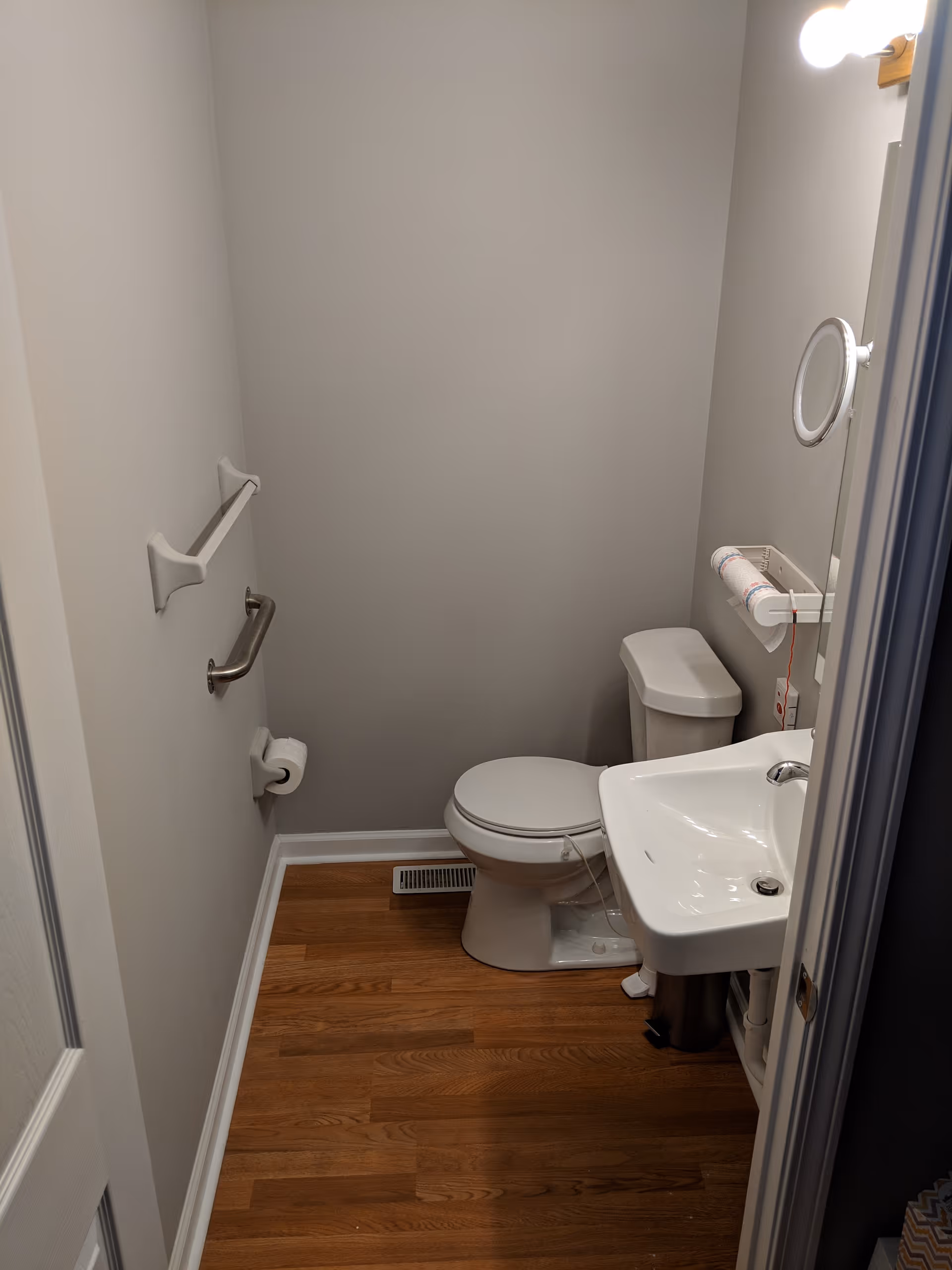 Small bathroom with a toilet and wall-mounted sink, grab bar and towel holder, and hardwood floor.