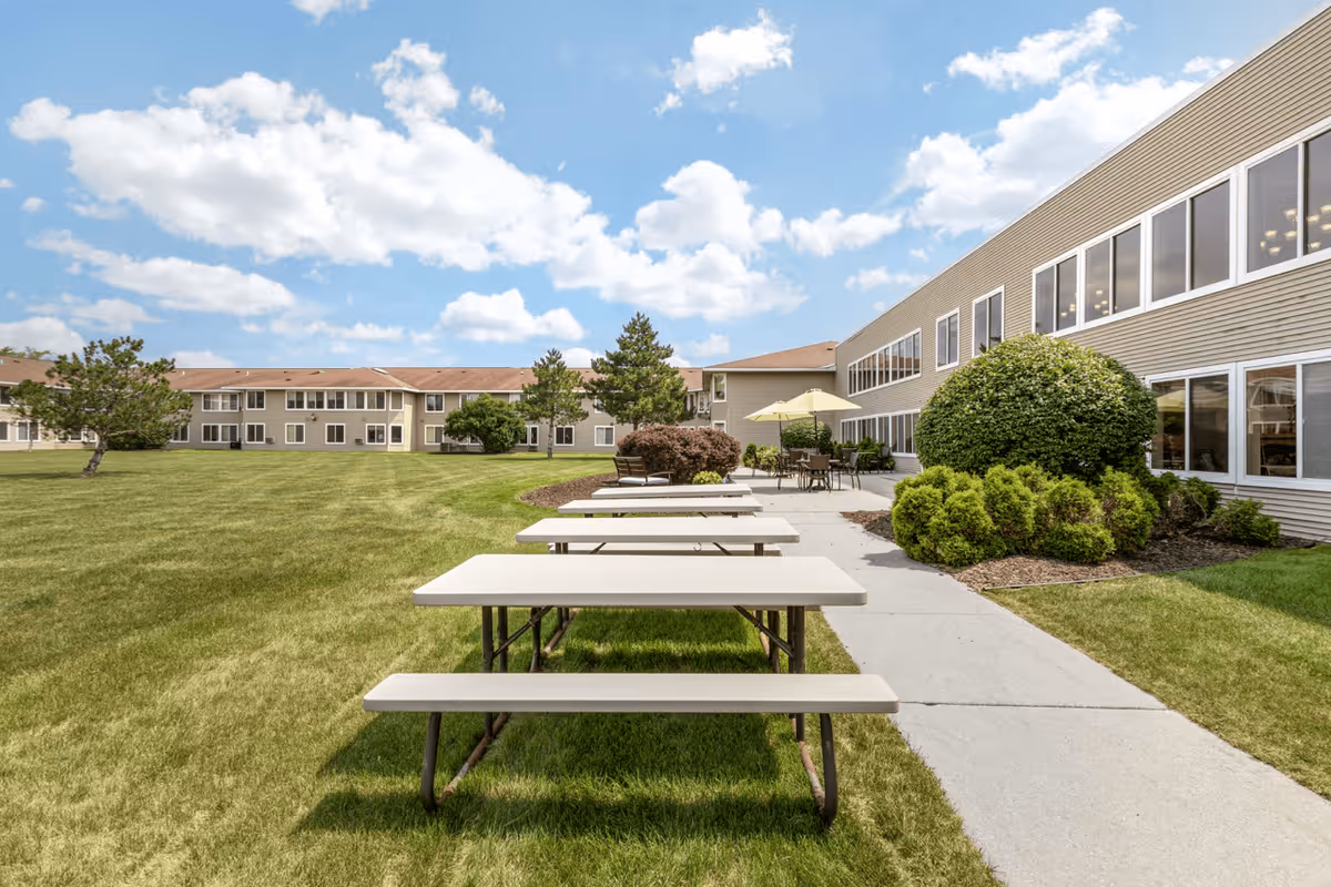 Outdoor area of Layton Terrace Senior Living featuring picnic tables with benches on a grassy lawn next to a paved walkway. The building has large windows and is surrounded by neatly trimmed bushes and trees under a partly cloudy blue sky.