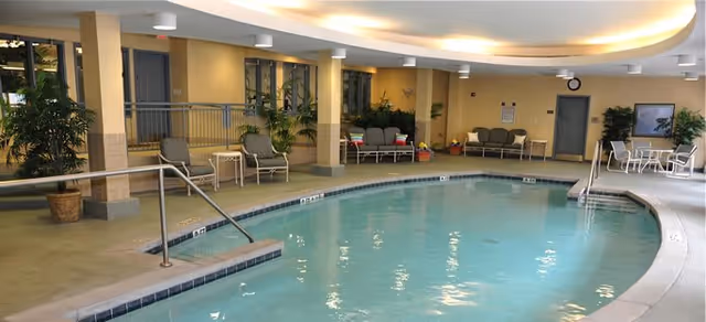 Indoor swimming pool area with clear water, surrounded by beige tiled flooring and several seating arrangements including chairs and small tables. Potted plants and large windows are visible along the walls, with soft overhead lighting illuminating the space.