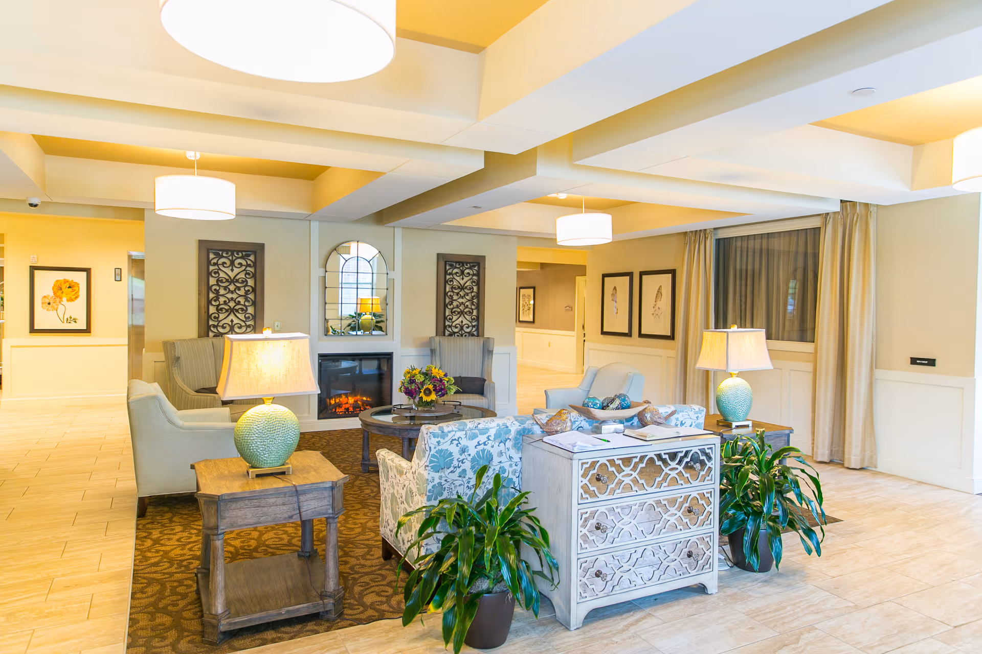A bright and welcoming senior living facility common area with comfortable armchairs and sofas arranged around a coffee table with a flower arrangement. There are two side tables with lamps, decorative wall art, a fireplace, and potted plants. The room has a tiled floor, beige walls, and large windows with curtains.