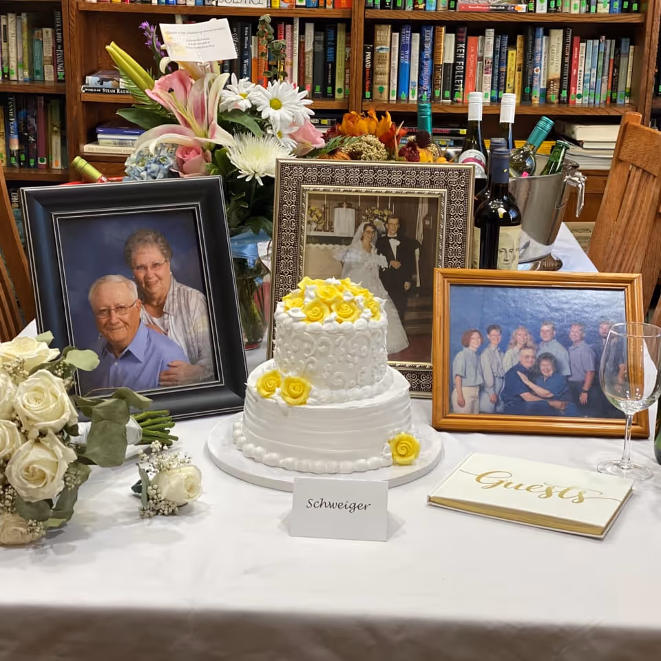 A table set with a white two-tier cake decorated with yellow roses, three framed photographs including a wedding photo, a couple portrait, and a family group photo, a bouquet of white roses, a guest book, and a bucket with bottles of wine. Behind the table is a bookshelf filled with books.