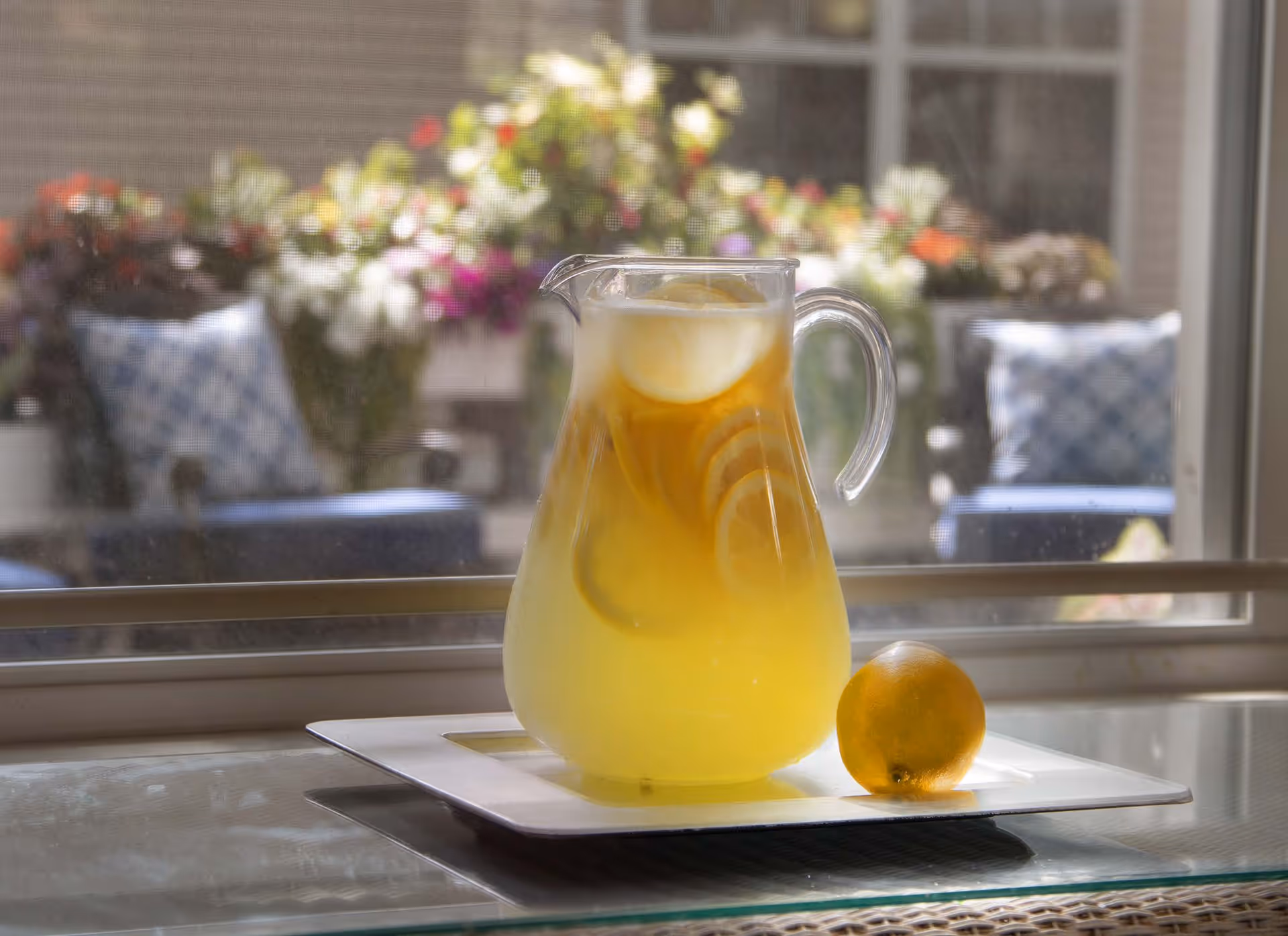 A glass pitcher filled with iced lemonade with lemon slices inside, placed on a white rectangular tray on a glass table. A whole lemon is also on the tray. In the background, there is a window showing an outdoor patio area with cushioned chairs and colorful flowers.