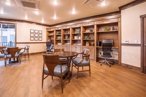 A well-lit interior room with wooden flooring and wooden furniture including round tables and chairs. There is a built-in wooden bookshelf along one wall with decorative plants, books, and a computer workstation with an office chair. The room has a warm and inviting atmosphere.
