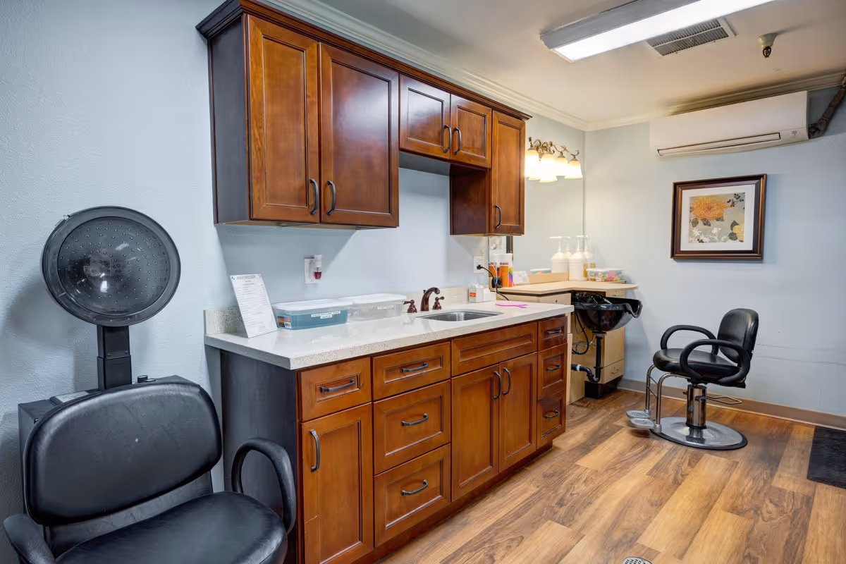 Interior view of a salon area in a senior living facility featuring two black salon chairs, a hair dryer hood, wooden cabinets with a countertop and sink, a small washing station, and a framed floral artwork on the wall.
