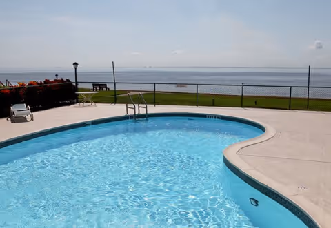 Outdoor swimming pool with clear blue water, surrounded by a concrete deck. In the background, there is a chain-link fence, green grass, a bench, and a view of a large body of water under a partly cloudy sky.