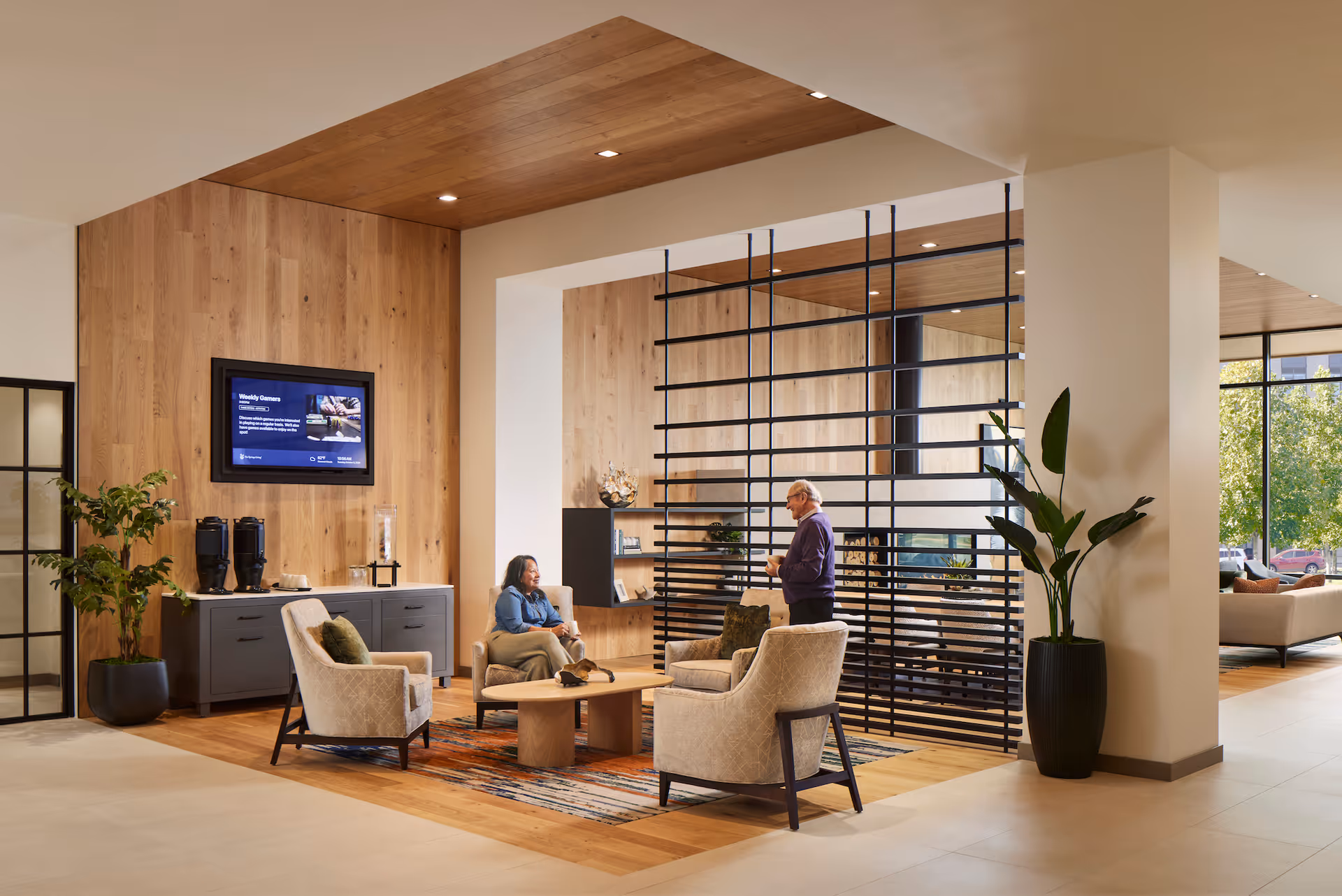 A modern and cozy common area in a senior living facility with wooden paneling on the walls and ceiling. The space features a seating arrangement with four cushioned armchairs around a wooden coffee table on a patterned rug. Two people are engaged in conversation, one seated and one standing. There are plants in large pots, a flat-screen TV mounted on the wall, and a decorative black metal grid partition separating the seating area from another part of the room with large windows showing greenery outside.