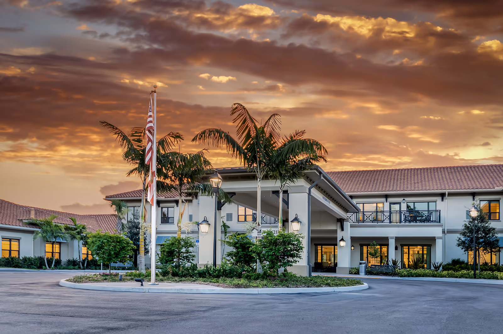 Exterior view of The Capstone at Royal Palm Senior Living facility at sunset, showing a two-story building with a covered entrance, palm trees, an American flag, and well-maintained landscaping.