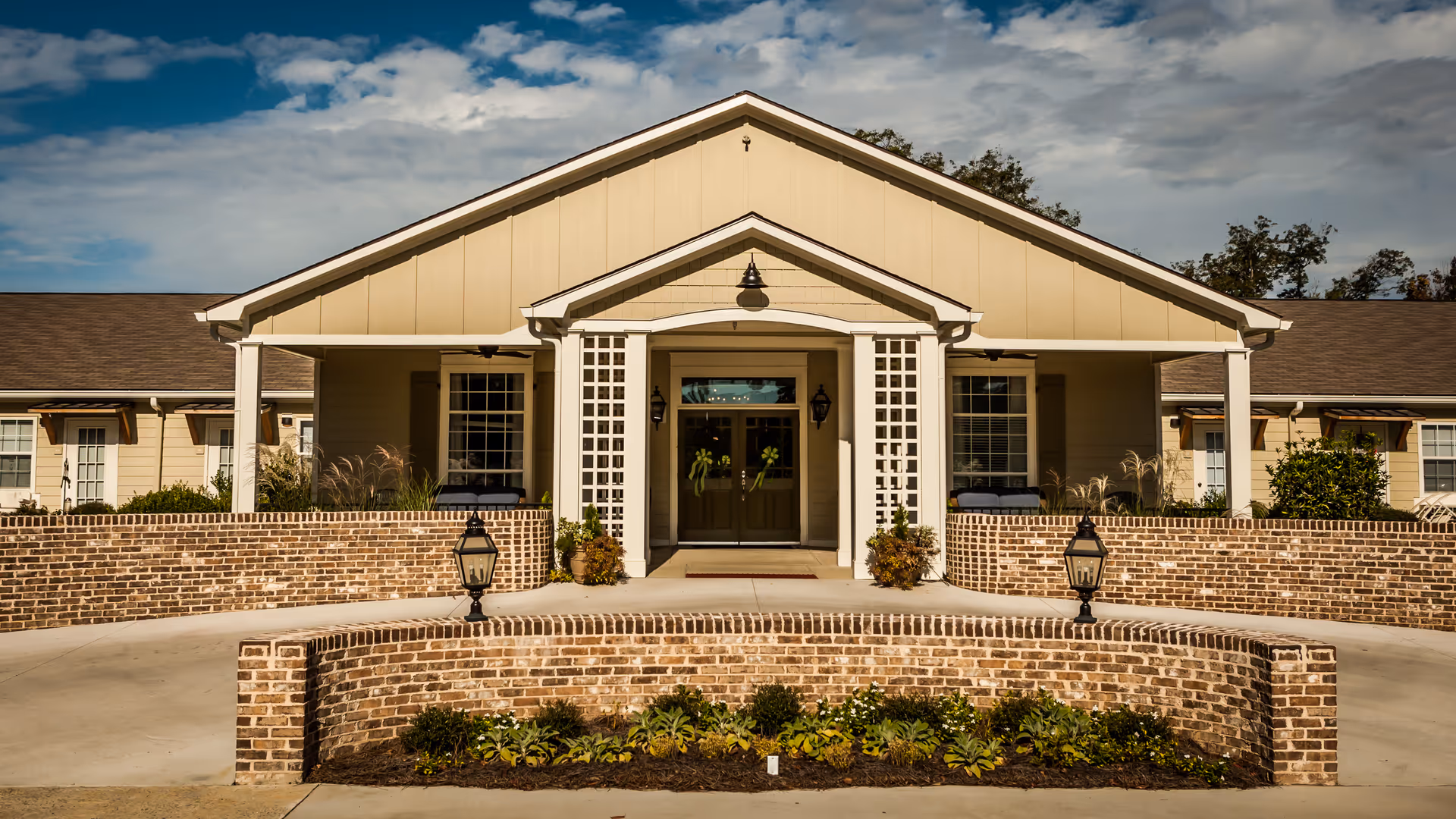 Front exterior view of Fellowship Cordelia Manor Assisted Living Community featuring a beige building with a peaked roof, brick walls, two lantern-style lights, and a landscaped garden bed in front under a partly cloudy sky.