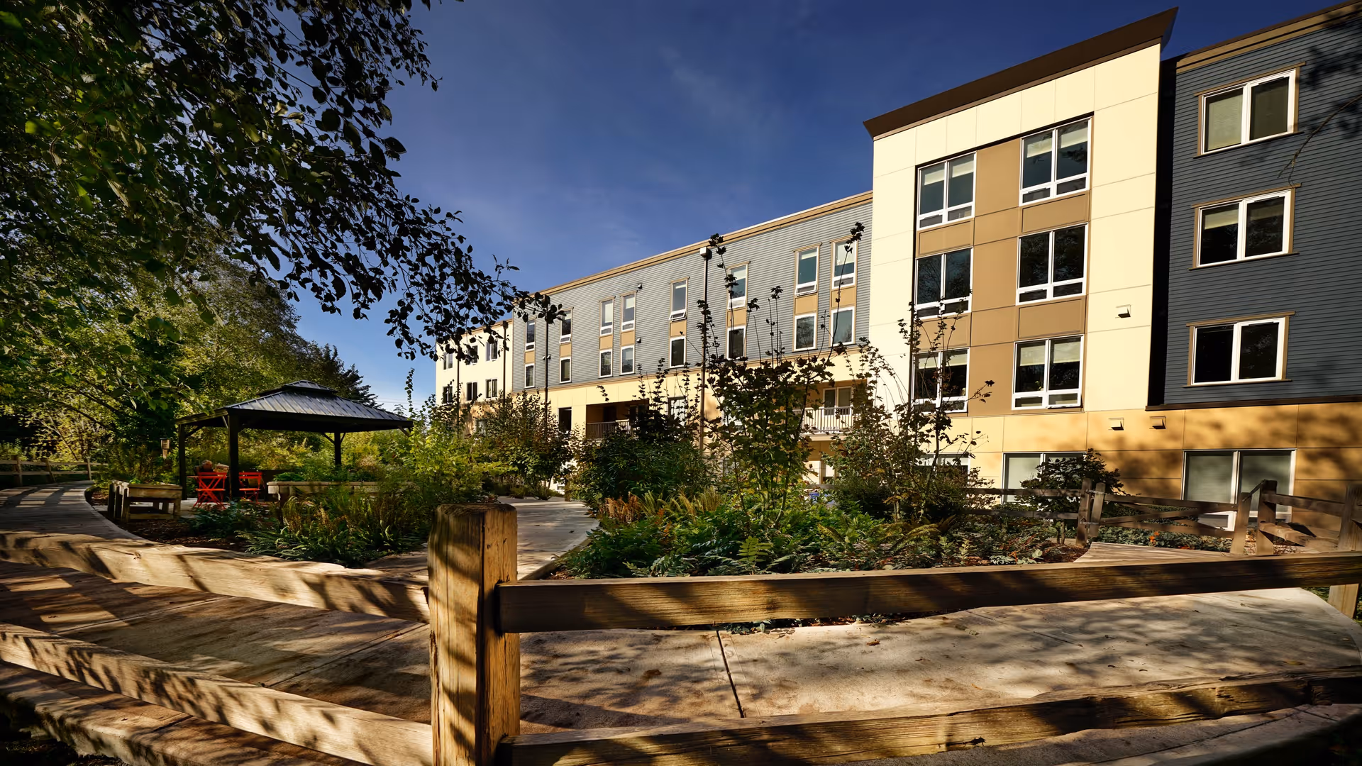 Outdoor view of Bainbridge Senior Living Madrona House showing a multi-story building with large windows, surrounded by landscaped gardens, trees, and a wooden fence. There is a gazebo with red chairs in the garden area under a clear blue sky.