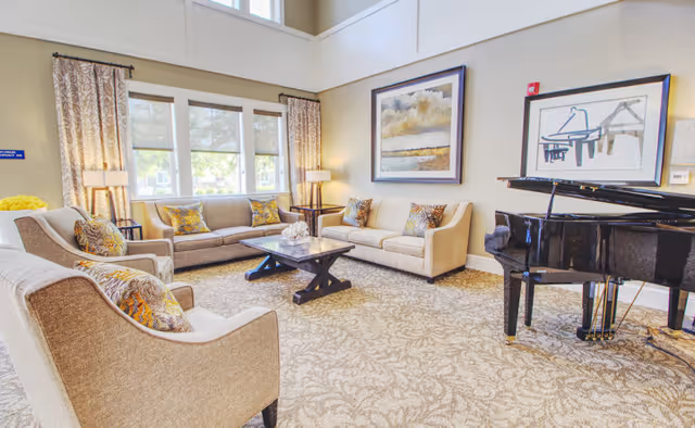 Bright communal living room with sofas, armchairs, a coffee table and a grand piano under tall windows.