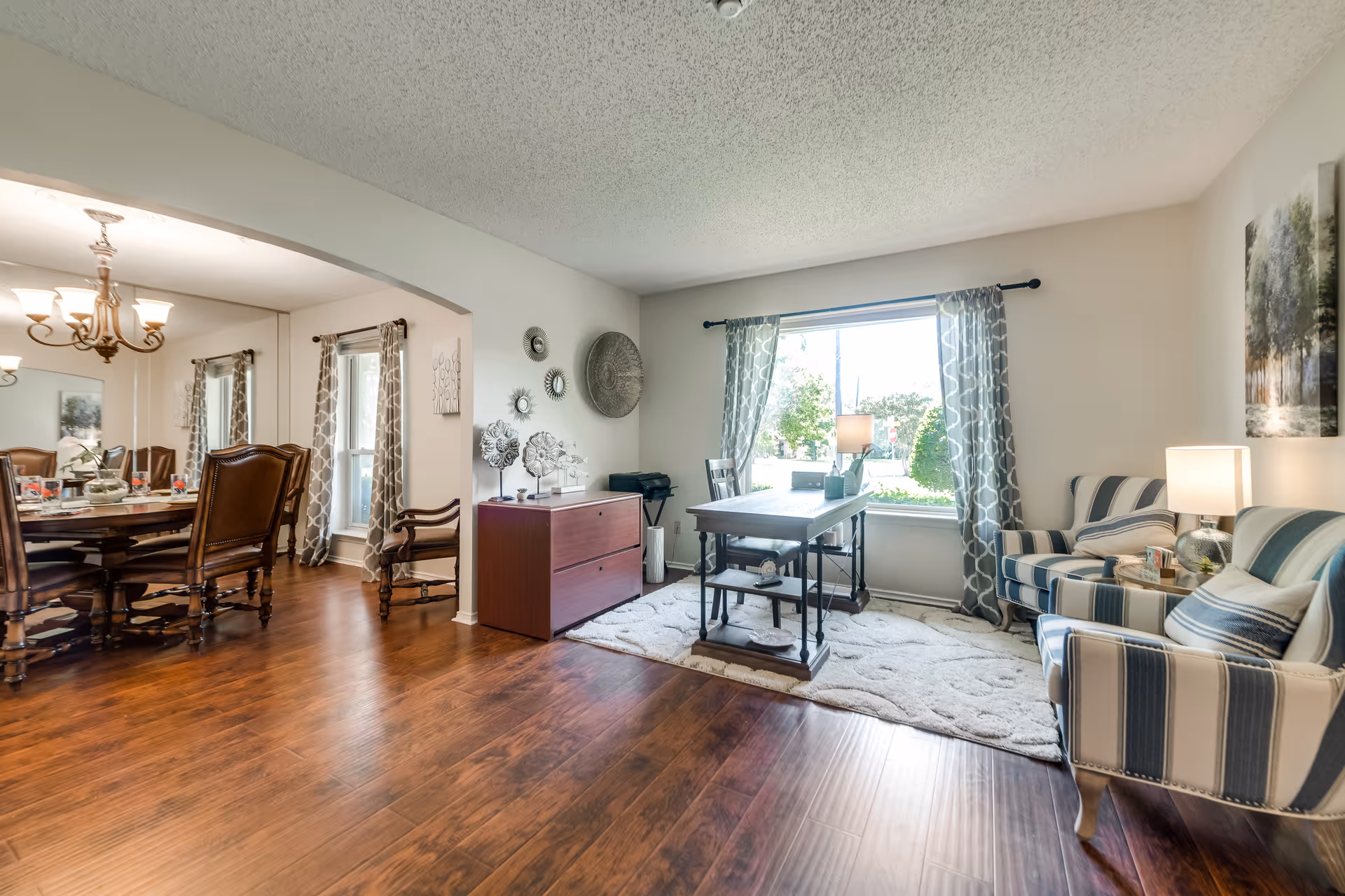 A bright and spacious interior room with wooden flooring featuring a sitting area with two striped armchairs, a small table with a lamp, and a large window with patterned curtains. Adjacent to this area is a dining room with a wooden dining table and chairs, a chandelier, and mirrored walls reflecting the room.