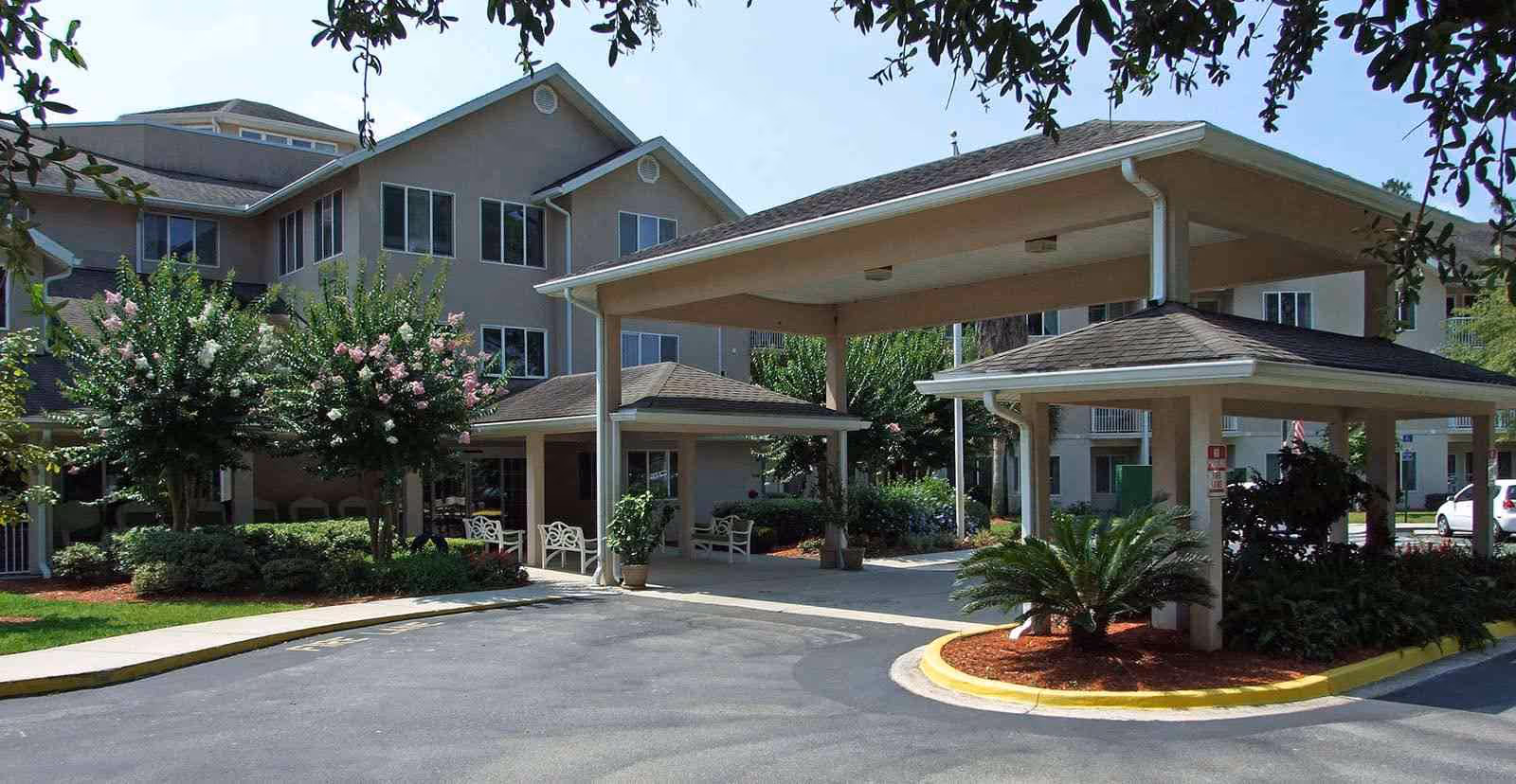 Exterior view of Augustine Landing senior living facility showing a covered entrance with multiple pillars, landscaped greenery including trees and shrubs, and a paved driveway.