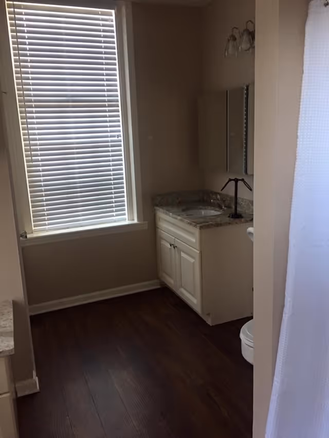 A bathroom with a window covered by white blinds, a white vanity with a granite countertop and a sink, a mirror above the sink, a light fixture with two bulbs, dark wood flooring, and a white shower curtain partially visible on the right side.