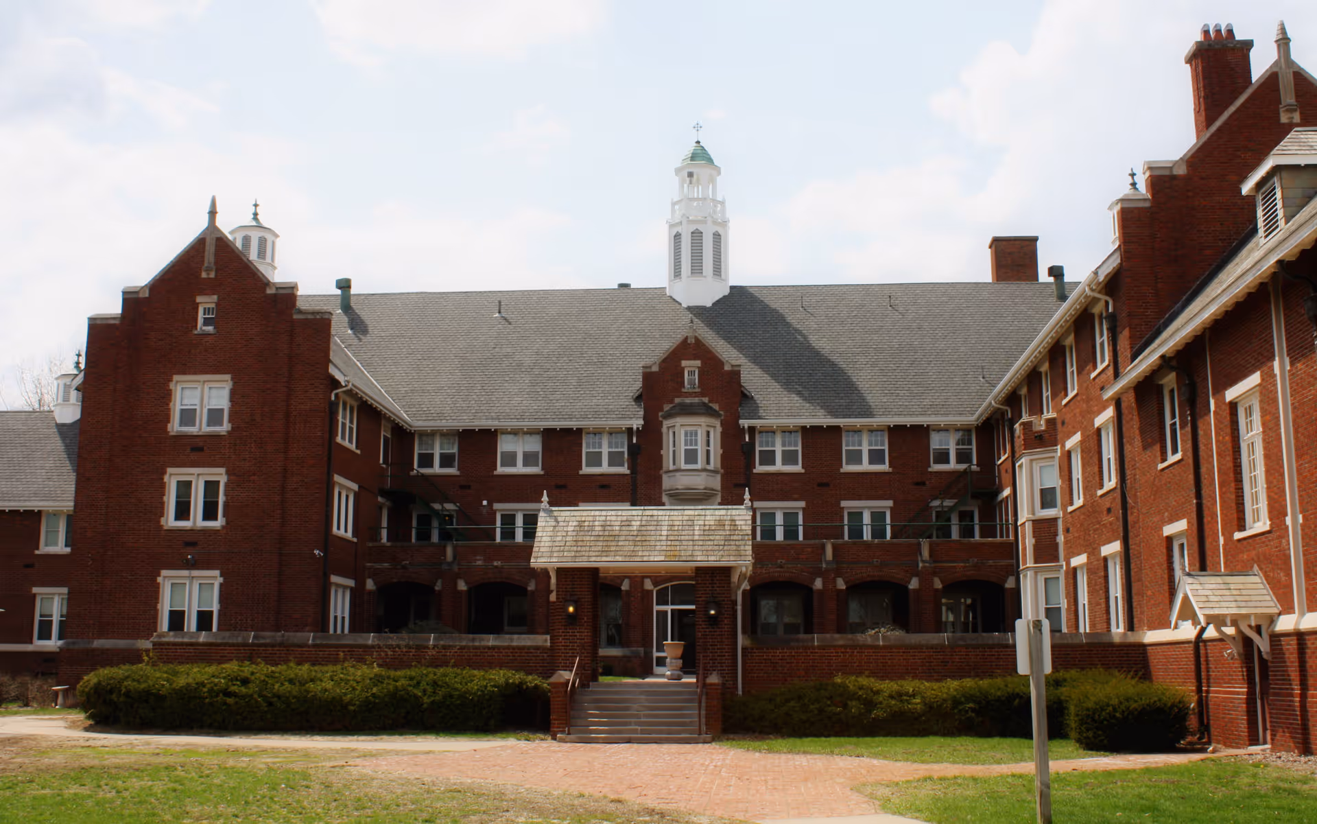 Exterior view of a large, multi-story brick building with a gray shingled roof and a central cupola. The building has multiple windows and a covered entrance with steps leading up to it. There is a grassy area and some bushes in front of the building under a partly cloudy sky.