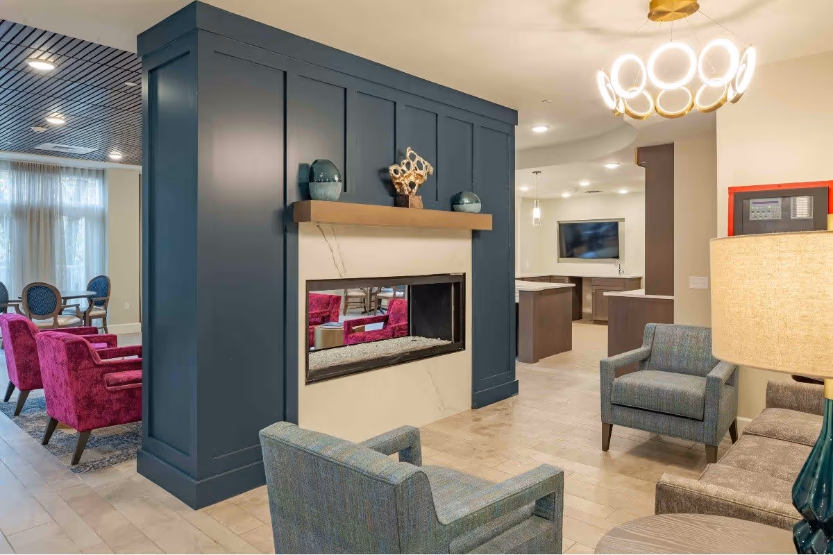A modern and cozy living room area in a senior living facility featuring a central double-sided fireplace with a dark blue paneled surround and a wooden mantel decorated with vases and a sculpture. The room includes comfortable seating with blue and purple upholstered chairs, a sofa, a round side table with a lamp, and a large flat-screen TV mounted on the wall in the background. The space is well-lit with ceiling lights and a stylish circular chandelier.