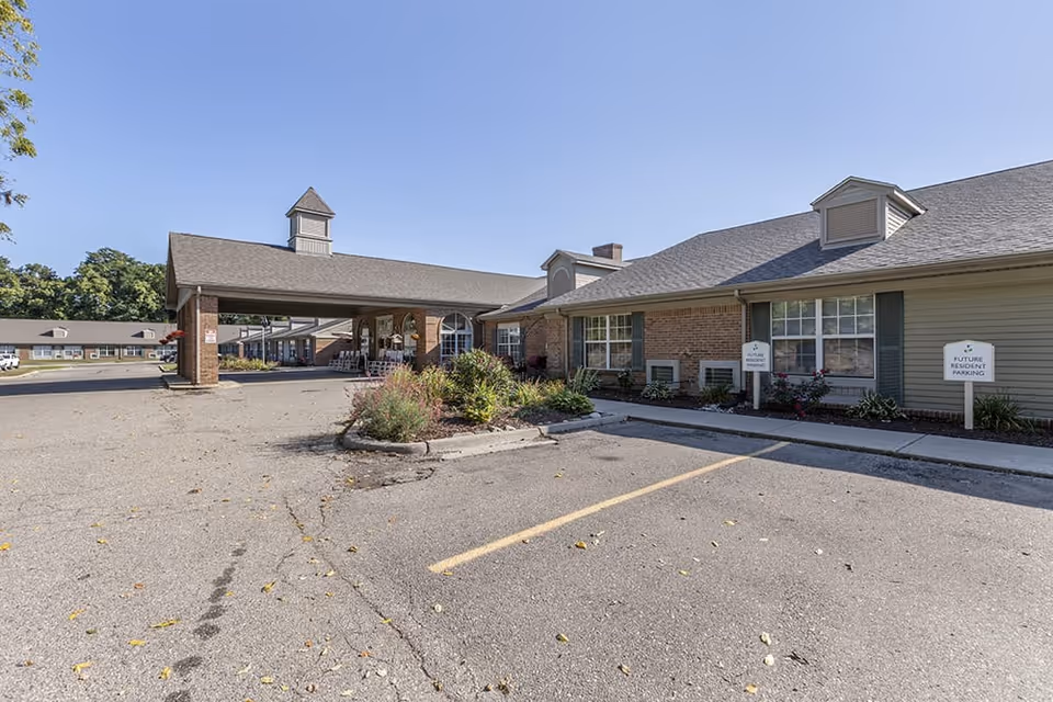 Exterior view of a senior living facility with a covered entrance, brick and siding walls, and a parking lot with signs indicating future resident parking. The sky is clear and blue.