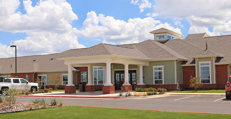 Exterior view of a single-story senior living facility building with a covered entrance supported by white columns, surrounded by a parking lot and landscaped greenery under a partly cloudy sky.