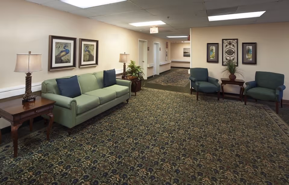 A hallway seating area in a senior living facility with a green sofa, two green armchairs, two wooden side tables with lamps, framed peacock artwork on the walls, and a patterned carpet covering the floor.