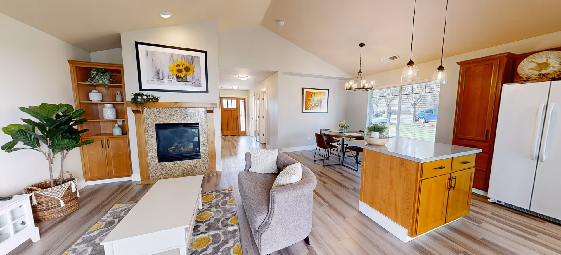 A bright and spacious open-concept living area featuring a cozy gray sofa with white pillows, a white coffee table on a patterned rug, a fireplace with a sunflower painting above it, built-in wooden shelves with decorative items, a kitchen island with wooden cabinetry and a white countertop, a dining table with four chairs near a large window, and a white refrigerator. The room has light wood flooring and neutral-colored walls.