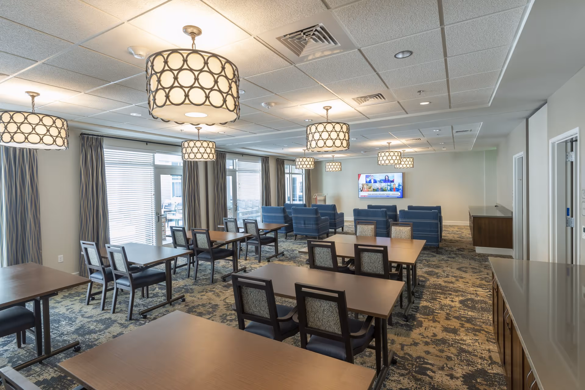 A spacious common area in a senior living facility with multiple tables and chairs arranged for dining or activities. The room features patterned carpet, large windows with curtains, and several modern circular pendant lights hanging from the ceiling. At the far end, there are blue upholstered sofas facing a wall-mounted flat-screen TV displaying a news program.