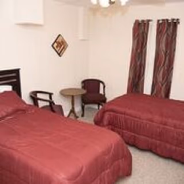 Small bedroom with two twin beds covered in red comforters, a round side table, two chairs, and matching curtains.