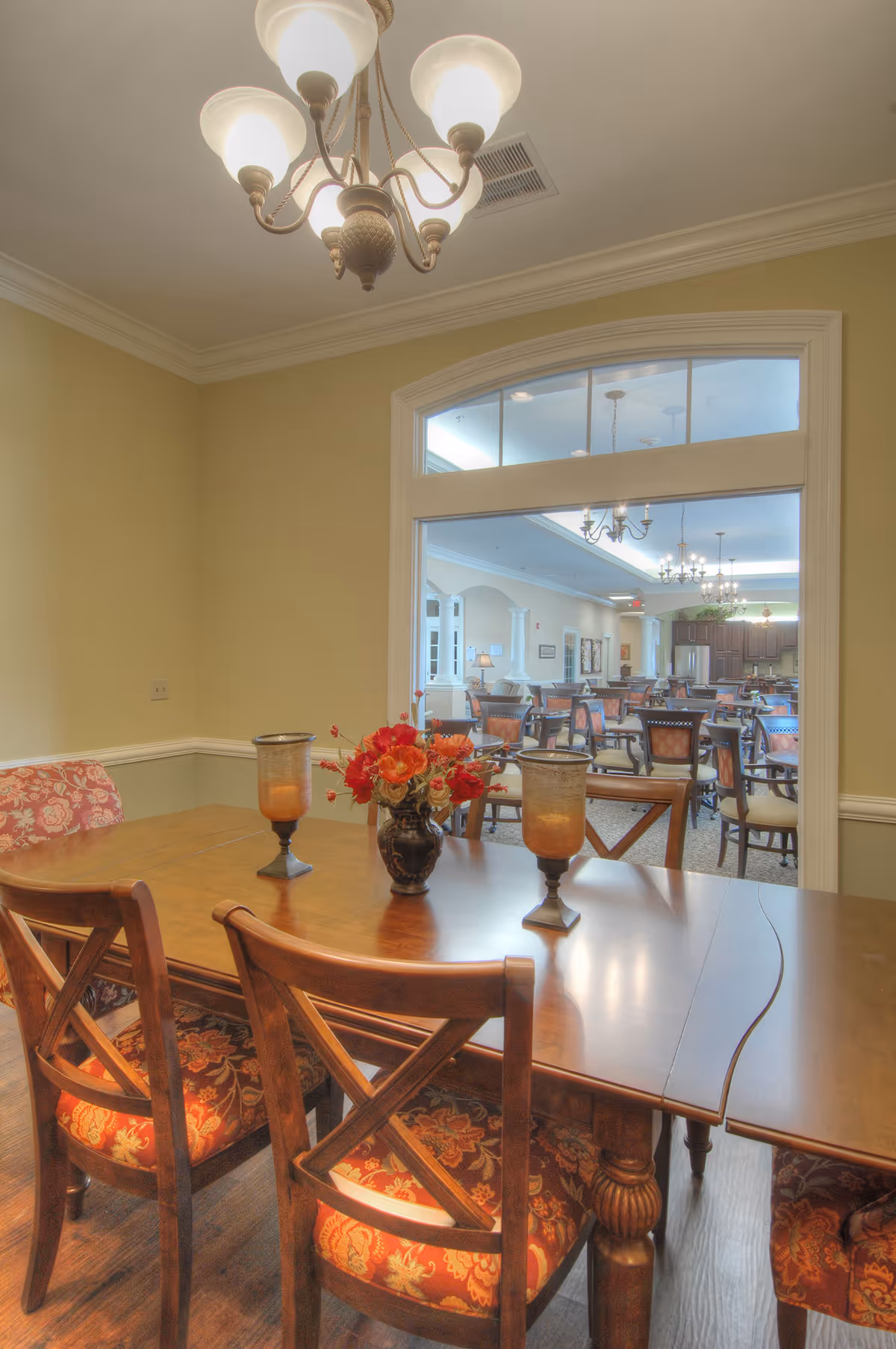 A dining area with a wooden table and four chairs with floral upholstery. On the table are two decorative candle holders and a vase with red and orange flowers. A chandelier with five lights hangs from the ceiling. Through a large window-like opening, another dining room with multiple tables and chairs is visible, featuring chandeliers and a kitchen area in the background.